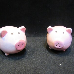 May include: Two ceramic pig figurines. The pigs are light pink with darker pink snouts and ears. They have small black eyes and are standing on four tiny legs. The background is black.