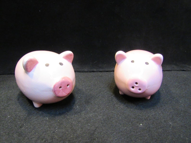 May include: Two ceramic pig figurines. The pigs are light pink with darker pink snouts and ears. They have small black eyes and are standing on four tiny legs. The background is black.