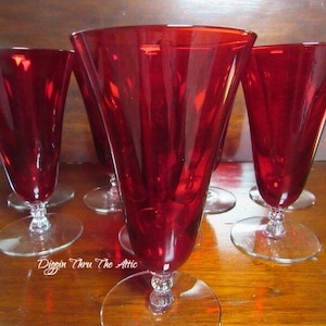 May include: Set of six red glass goblets with a clear stem and base. The goblets are arranged in a semi-circle on a wooden surface.