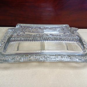 Puede incluir: Una mantequera de vidrio transparente con forma rectangular y detalles ornamentados y en relieve alrededor de los bordes y el asa. El plato tiene tapa y está sobre una superficie clara.