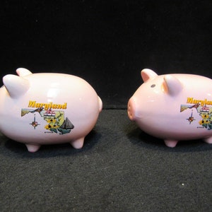 May include: Two pink ceramic piggy banks. Each bank has a map of Maryland with the word "Maryland" in yellow. The banks have small ears, a snout, and four small legs. The banks are on a black background.