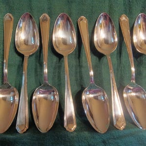 Puede incluir: Colección de siete cucharas plateadas con una superficie pulida y reflectante. Las cucharas tienen un diseño clásico con cuencos alargados y mangos decorativos. Se disponen sobre un paño verde, mostrando su elegante forma.