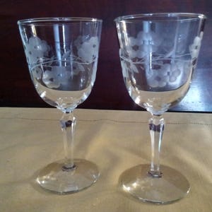 May include: Two clear wine glasses with etched floral designs. The glasses have a rounded bowl, a slender stem, and a circular base. The floral pattern is etched around the top of the bowl. The glasses are displayed on a light yellow surface.
