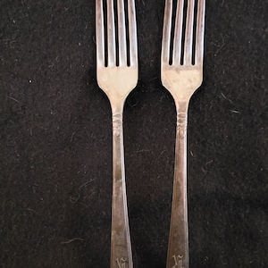 May include: Two silver forks with four tines each, arranged vertically against a dark background. The handles are engraved with an ornate initial design. The forks are likely vintage or antique.