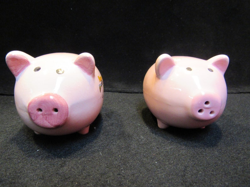 May include: Two pink ceramic pig figurines. Each pig has a pink snout with nostrils, small ears, and tiny black eyes. The pigs are on a dark surface.