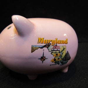 May include: A light pink ceramic piggy bank with a Maryland design. The bank features a map of Maryland with a bird, flowers, and a sailboat. The word "Maryland" is printed in yellow.
