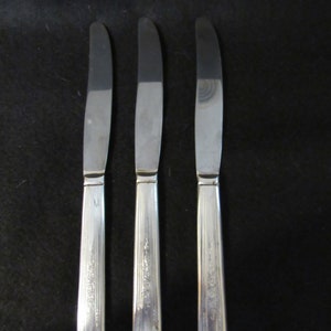 May include: Three silver knives with a polished blade and ornate handles. The handles feature a vertical, ridged pattern with floral detailing. The knives are displayed vertically against a dark backdrop.