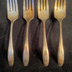 May include: Four vintage silver forks with tines and long handles. Each fork has a decorative initial engraved on the handle. The forks are arranged in a row against a dark background. The silver has a slight patina.