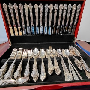 May include: A silver-toned flatware set arranged in a dark wood storage chest with a black velvet interior. The collection includes multiple knives standing upright in the lid, with various forks and spoons organized in the base slots.
