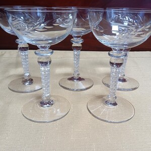 May include: A set of five clear crystal champagne coupe glasses. Each glass features a round base, a textured stem, and a bowl etched with a leaf design. The glasses are arranged on a light beige surface.