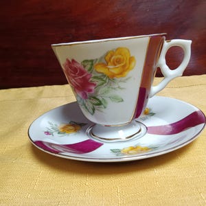 May include: A vintage teacup and saucer set. The white porcelain cup features a floral design with pink and yellow roses, accented with green leaves. The saucer mirrors the design with a magenta stripe. The cup has a gold trim.
