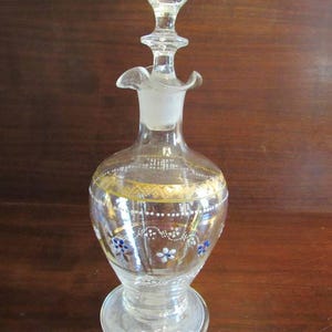 May include: A clear glass bottle with a gold and white floral design. The bottle has a stopper with a round top and a ruffled edge.