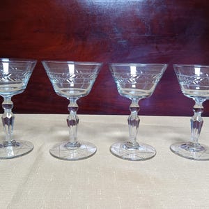 Puede incluir: Cuatro copas de champán transparentes vintage. Cada copa presenta un diseño grabado decorativo alrededor del cuenco y en la base. Los tallos tienen un diseño facetado. Las copas están dispuestas sobre una superficie clara.