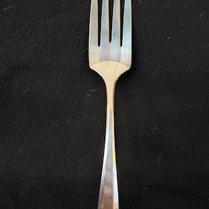 May include: A silver-toned metal fork featuring four long, slender tines and a wide, flat neck that transitions into a smooth, tapered handle. The handle displays a small, engraved cursive letter at the base, reflecting a polished, metallic finish.