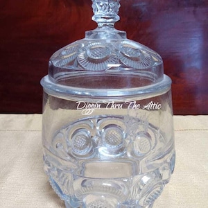 May include: Clear glass jar with a lid. The jar has a decorative pattern of circles and floral designs. The lid has a faceted knob. The text "Diggin Thru The Attic" is visible on the jar.