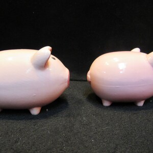 May include: Two pink ceramic piggy banks. One pig is facing left, showing its snout, ear, and eye. The other pig is facing right, showing its ear and rear. Both pigs have four small legs.
