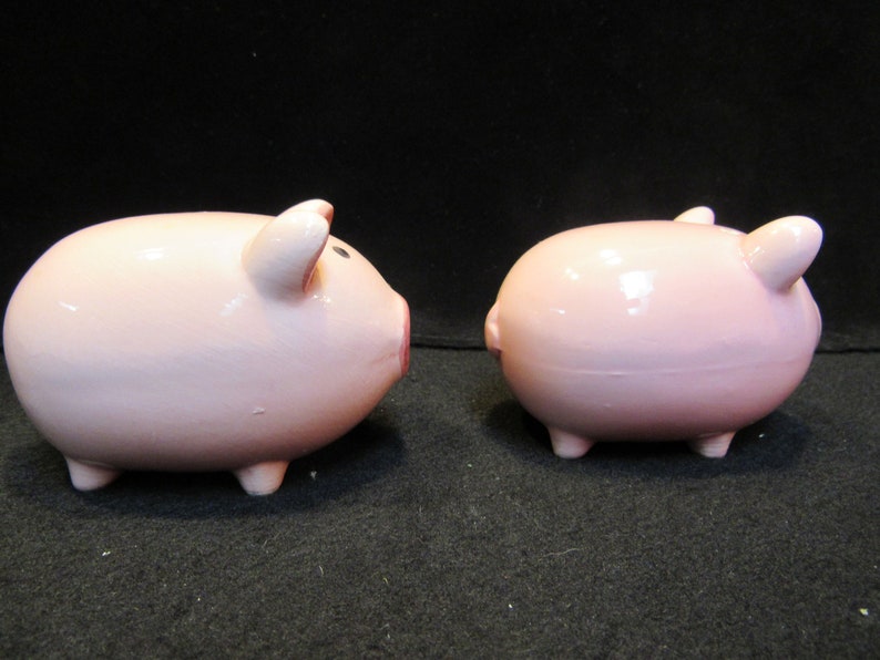 May include: Two pink ceramic piggy banks. One pig is facing left, showing its snout, ear, and eye. The other pig is facing right, showing its ear and rear. Both pigs have four small legs.