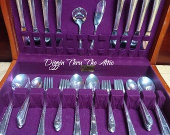 May include: A vintage silverware set displayed in a wooden case with a purple velvet lining. The set includes forks, knives, spoons, and serving utensils. The text "Diggin Thru The Attic" is visible on the bottom of the image.