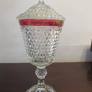 Puede incluir: Un plat de cristal transparente para dulces con un patrón de diamante y una banda roja alrededor de la parte superior. El plato tiene una tapa y se asienta sobre una base de pedestal.