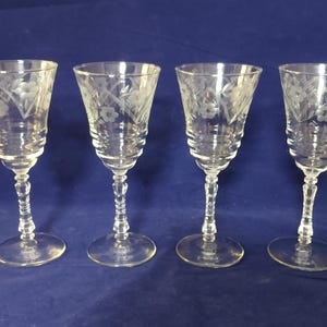 May include: A set of four clear glass goblets featuring an etched floral pattern around the upper rim. Each glass has a multi-tiered, ribbed bowl design that tapers down to a faceted stem and a circular base, providing a consistent look.