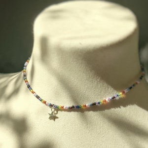 Könnte beinhalten: Ein mit bunten Perlen besetztes Choker-Halsband. Die Halskette hat kleine, farbige Perlen und einen silbernen Sternanhänger. Die Halskette wird auf einer Schaufensterpuppe präsentiert.