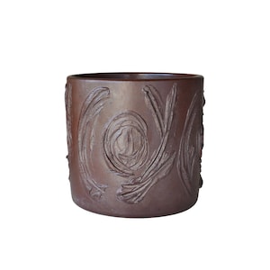 May include: A brown ceramic planter with a cylindrical shape. The exterior features a carved floral design. The planter is suitable for indoor or outdoor use and adds a touch of rustic charm to any space.