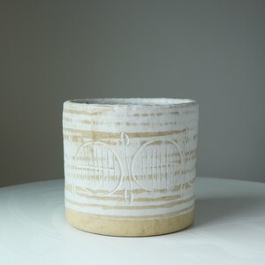 May include: White ceramic planter with a textured design of horizontal lines and circles. The planter has a light brown base.