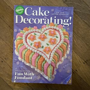 May include: A copy of the 2005 Wilton Yearbook, "Cake Decorating!" features a heart-shaped cake with orange flower decorations and a white pearl border. The cover also includes the text "Fun With Fondant."