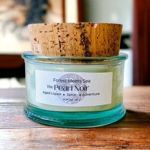 May include: A glass jar candle with a cork lid. The label reads "Forest Meets Sea The Pearl Noir Aged Liquor Spice Adventure NT WT 9 oz/255 g".