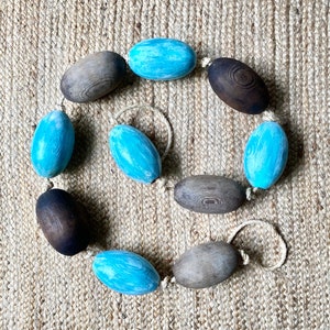 May include: A string of wooden and blue painted oval-shaped ornaments, connected with twine, on a woven jute background.