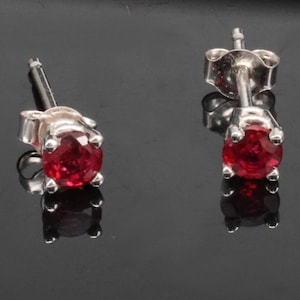 May include: A pair of stud earrings featuring round, faceted red stones set in silver-toned metal prongs. Each earring includes a post back mechanism, showcasing a clear, brilliant red colour and a polished metallic finish on the setting.