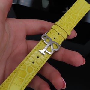 May include: A bright yellow watch strap with a textured, crocodile-like pattern. The strap features a silver-colored buckle with an abstract design. The strap is held in a hand against a dark background.