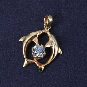 May include: A gold pendant featuring two dolphins facing each other in a circular formation, with a single round, clear, faceted gemstone set in the center. The pendant includes a small gold bail at the top for attachment to a chain or necklace.