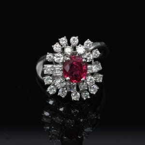 May include: A silver ring with a central oval ruby, encircled by a halo of round and baguette-cut diamonds. The ring is set against a black backdrop, accentuating the brilliance of the gemstones. The ring's design is elegant.