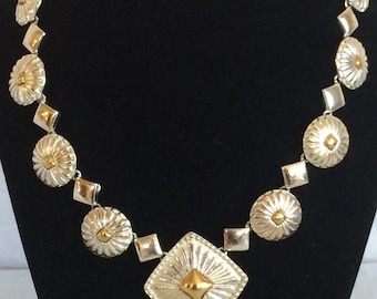 May include: A silver and gold necklace with a large square pendant. The necklace is made of a chain with round and square links. The pendant has a textured surface and a small gold square in the center.