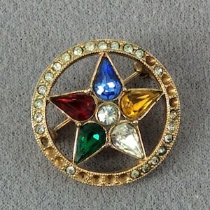 Puede incluir: Un broche dorado con un diseño de estrella de cinco puntas. La estrella está engastada con cinco piedras facetadas: roja, azul, verde, amarilla y transparente. La estrella está rodeada por un círculo de piedras transparentes más pequeñas.