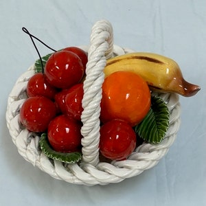 Könnte beinhalten: Ein weißer Keramik-Obstkorb mit einem Seilgriff. Der Korb ist gefüllt mit roten Kirschen, einer gelben Banane, einer orangen Orange und grünen Blättern.