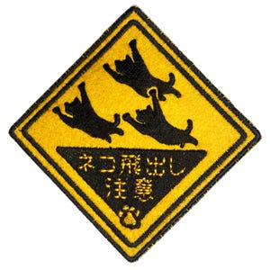 以下が含まれることがあります： 黄色いひし形の刺繍パッチ、黒い縁取り。3匹の黒い猫のシルエットが遊び心のあるポーズで描かれ、日本語のテキストと足跡が特徴です。テキストは「ネコ飛び出し注意」と書かれています。