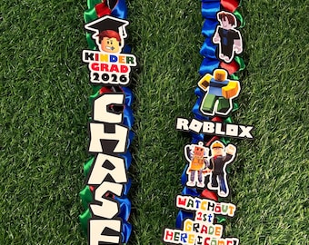 May include: A graduation lei made of braided red, green, and blue satin ribbons. It features cutouts of a character in a graduation cap with the text KINDER GRAD 2026, the name CHASE, Roblox characters, the Roblox logo, and WATCH OUT 1ST GRADE HERE I COME.