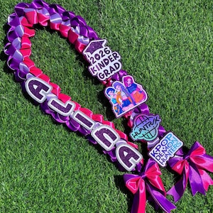 May include: A braided lei in shades of purple, pink, and white, with the name "ALIA" spelled out in large, glittery letters. It features decorative charms with the text "2026 Kinder Grad" and "KPOP DEMON HUNTERS".