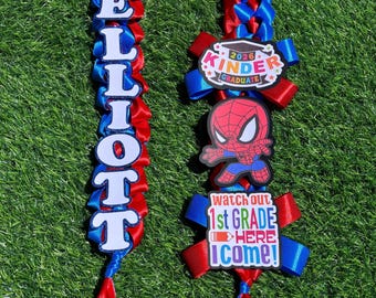 May include: A red, white, and blue graduation lei with the name "ELLIOTT" spelled out in white letters. The lei is decorated with a "2026 Kinder Graduate" emblem, a Spider-Man graphic, and a "Watch out 1st Grade HERE I COME!" emblem.