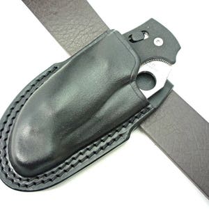 May include: A black leather belt holster with a stitched edge, designed to hold a folding knife. The knife is partially visible in the holster, with a black handle and a silver blade.