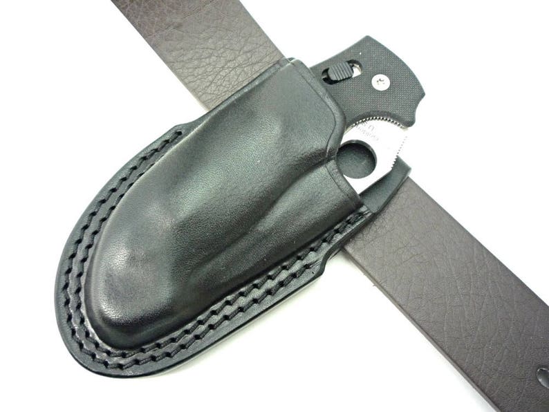 May include: A black leather belt holster with a stitched edge, designed to hold a folding knife. The knife is partially visible in the holster, with a black handle and a silver blade.