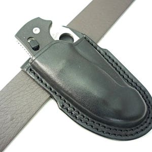 May include: A black leather knife sheath with a stitched edge. The sheath is designed to fit a folding knife with a clip. The sheath is attached to a brown leather belt.