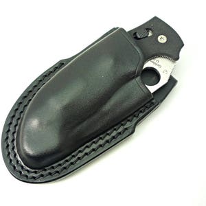 May include: A black leather knife sheath with a stitched edge. The sheath is designed to hold a folding pocket knife with a partial blade exposed.