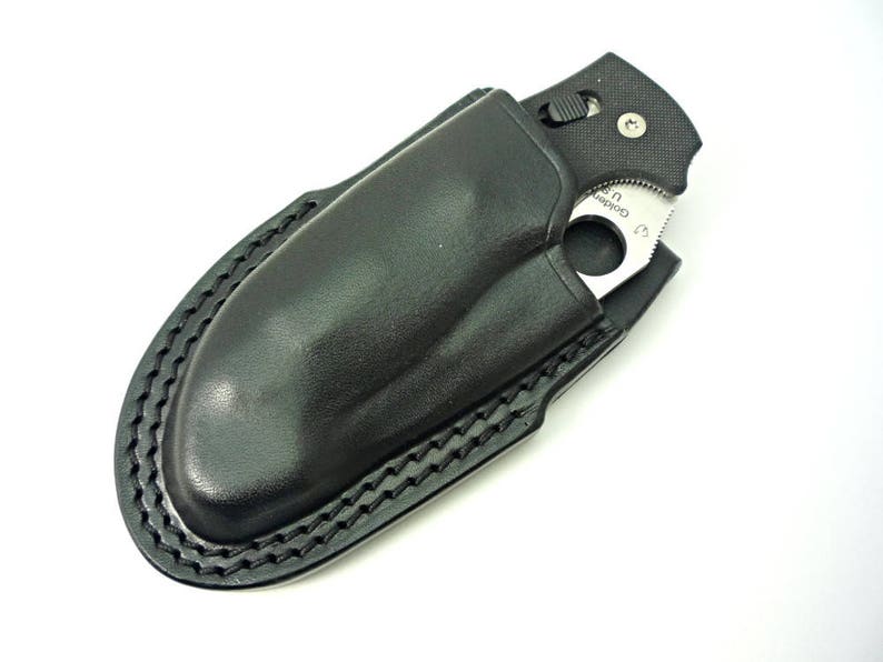 May include: A black leather knife sheath with a stitched edge. The sheath is designed to hold a folding pocket knife with a partial blade exposed.