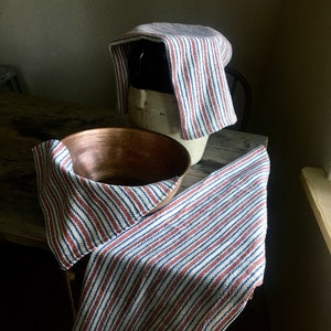 May include: Two striped kitchen towels, one covering a ceramic crock and the other draped over a copper bowl. The towels are white with red and blue stripes.