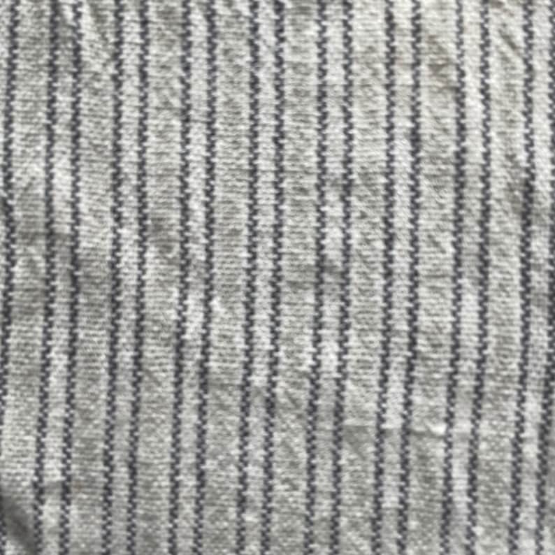 May include: Close-up of a white fabric with thin, vertical, dark gray stripes. The fabric has a textured, woven appearance.