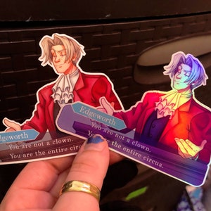 May include: Two holographic stickers featuring a cartoon character with the text "Edgeworth You are not a clown. You are the entire circus."