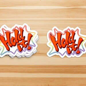 May include: A stack of "Hold It!" stickers with a rainbow border. The text is in a bold, orange font.